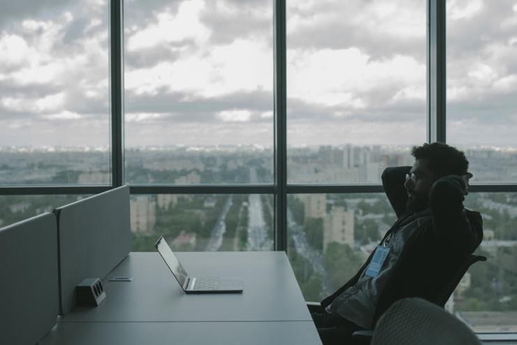 Man sitting in an office looking at his laptop with a view of the city from his window